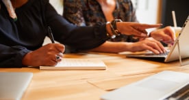 Two women sitting at a desk taking notes on a laptop and notepad